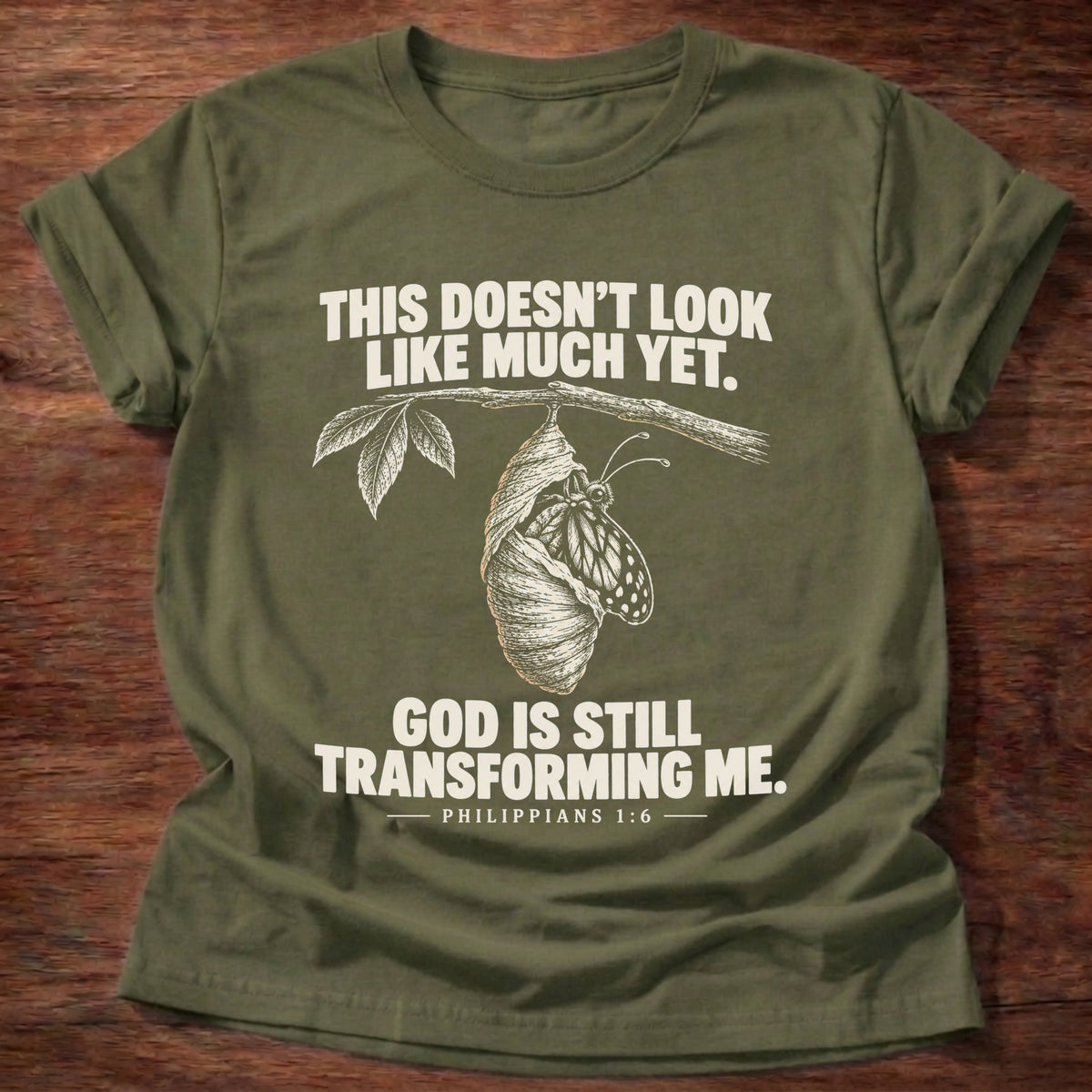 Military Green t-shirt printed with a chrysalis hanging from a branch graphic and 'This doesn't look like much yet. God is still transforming me.' Philippians 1:6 faith based gift