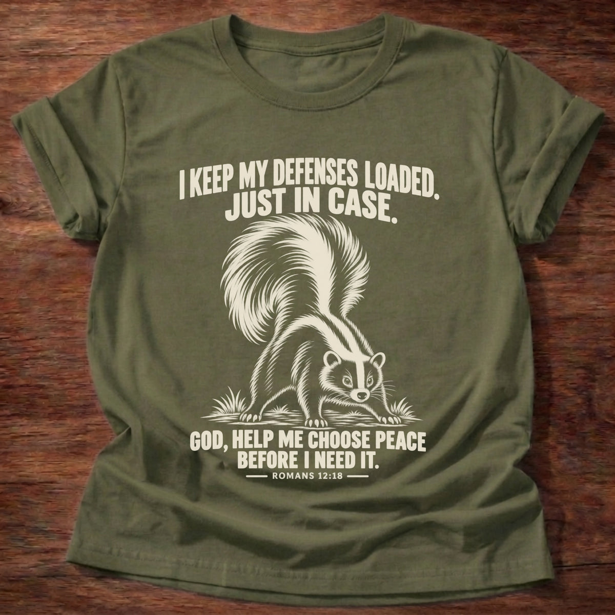 Military Green t-shirt printed with a skunk with its tail raised, ready to spray graphic and 'I keep my defenses loaded. Just in case. God, help me choose peace before I need it.' Romans 12:18 faith based gift