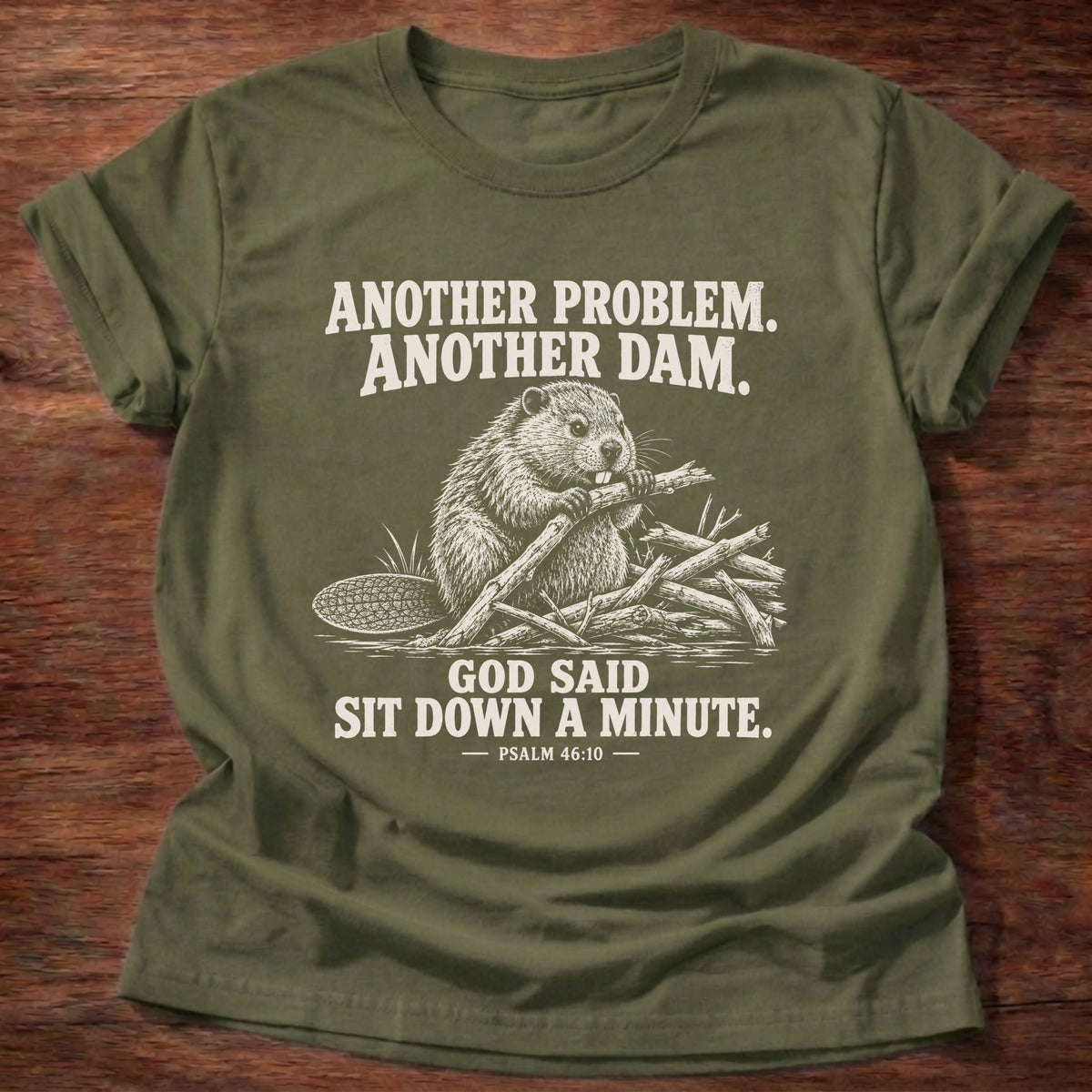 Military Green t-shirt printed with a beaver mid-build with another branch graphic and 'Another problem. Another dam. God said sit down a minute.' Psalm 46:10 faith based gift