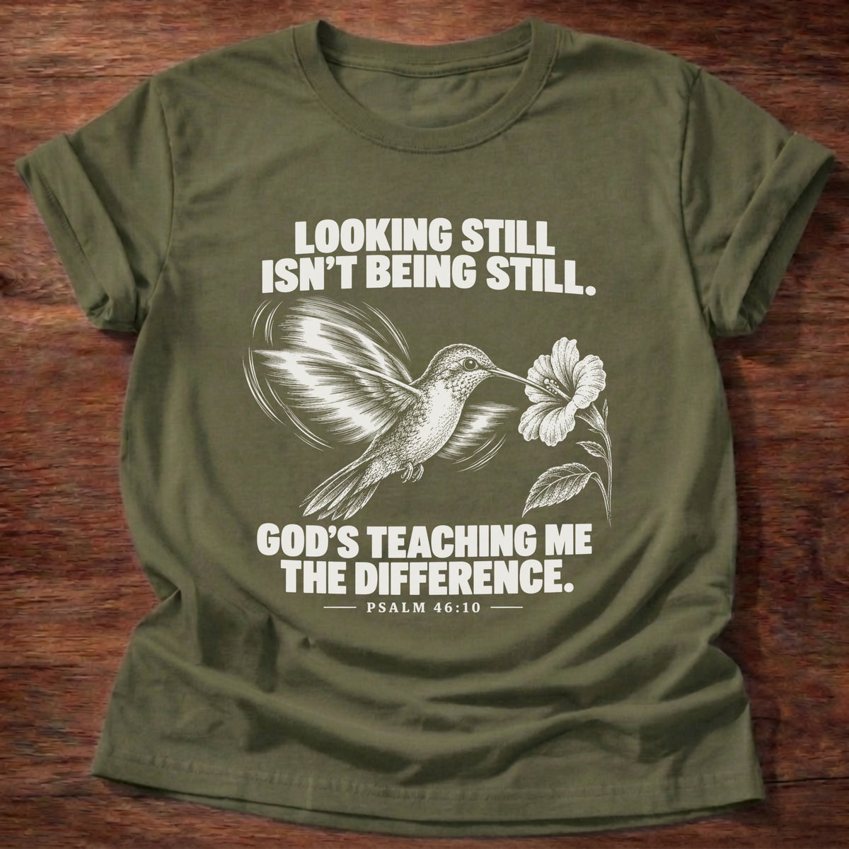 Military Green t-shirt printed with a hummingbird hovering beside a flower graphic and 'Looking still isn't being still. God's teaching me the difference.' Psalm 46:10 faith based gift