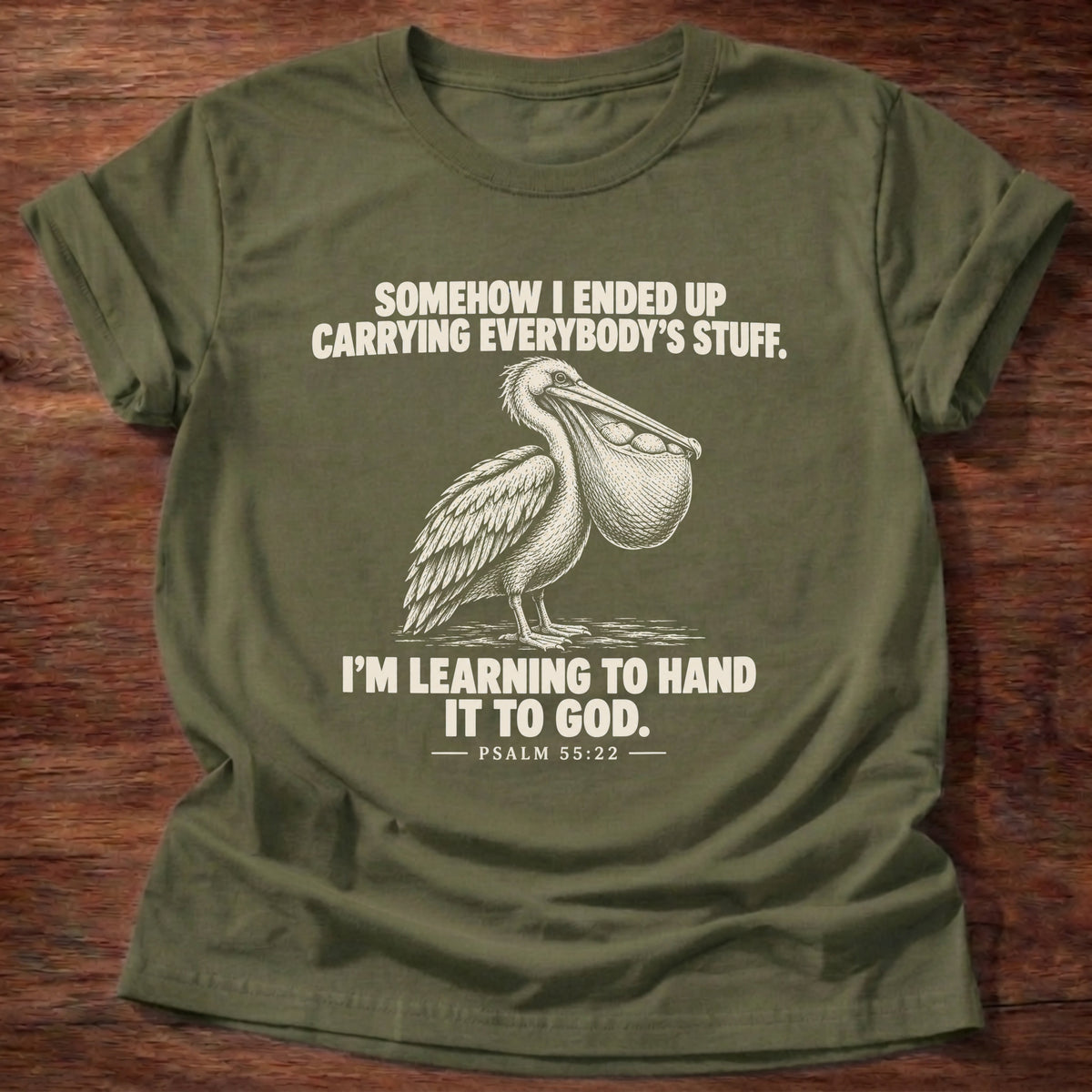 Military Green t-shirt printed with a pelican with a pouch full of fish graphic and 'Somehow I ended up carrying everybody's stuff. I'm learning to hand it to God.' Psalm 55:22 faith based gift