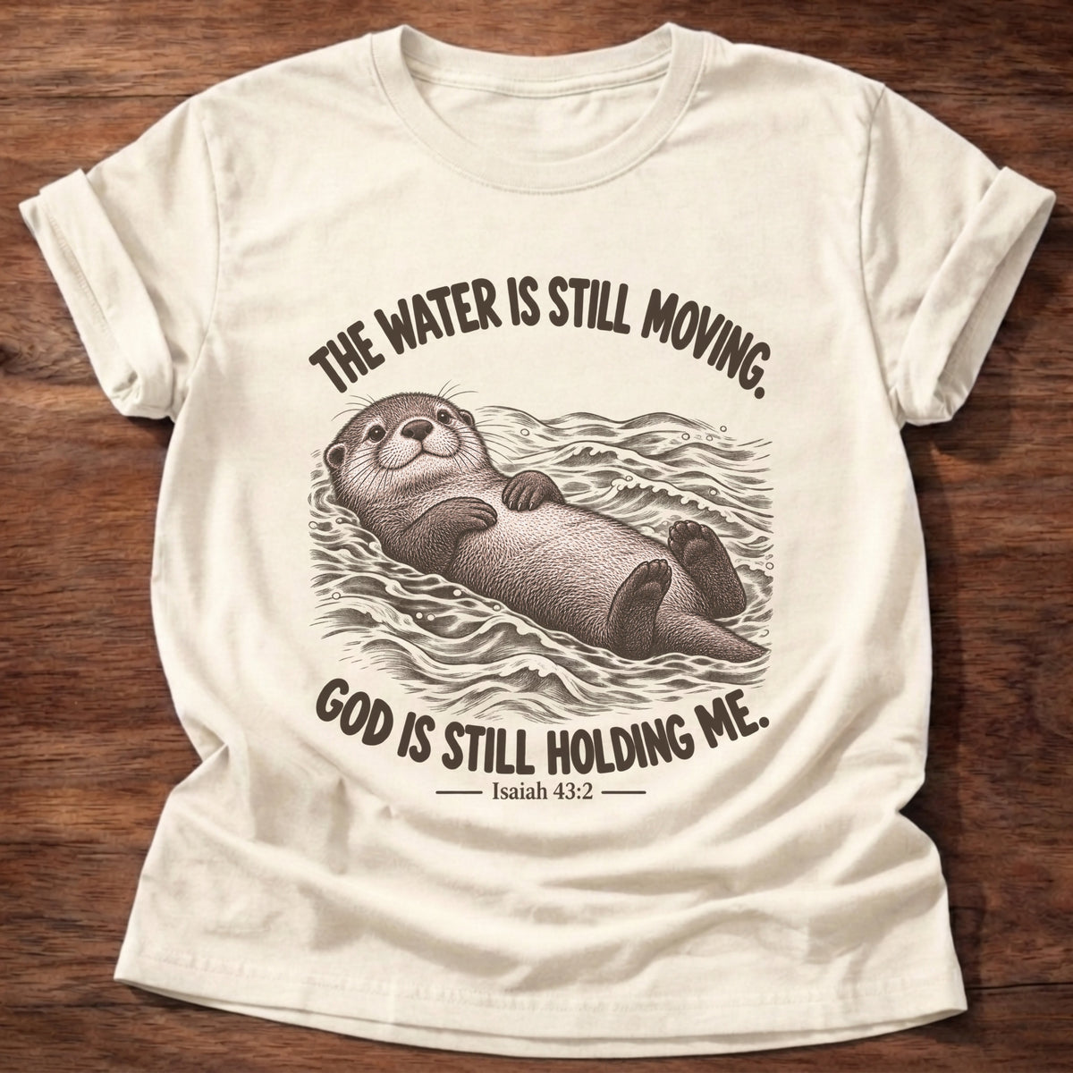 Natural t-shirt printed with an otter floating on its back in moving water graphic and “The water is still moving. God is still holding me.” Isaiah 43:2 faith based gift