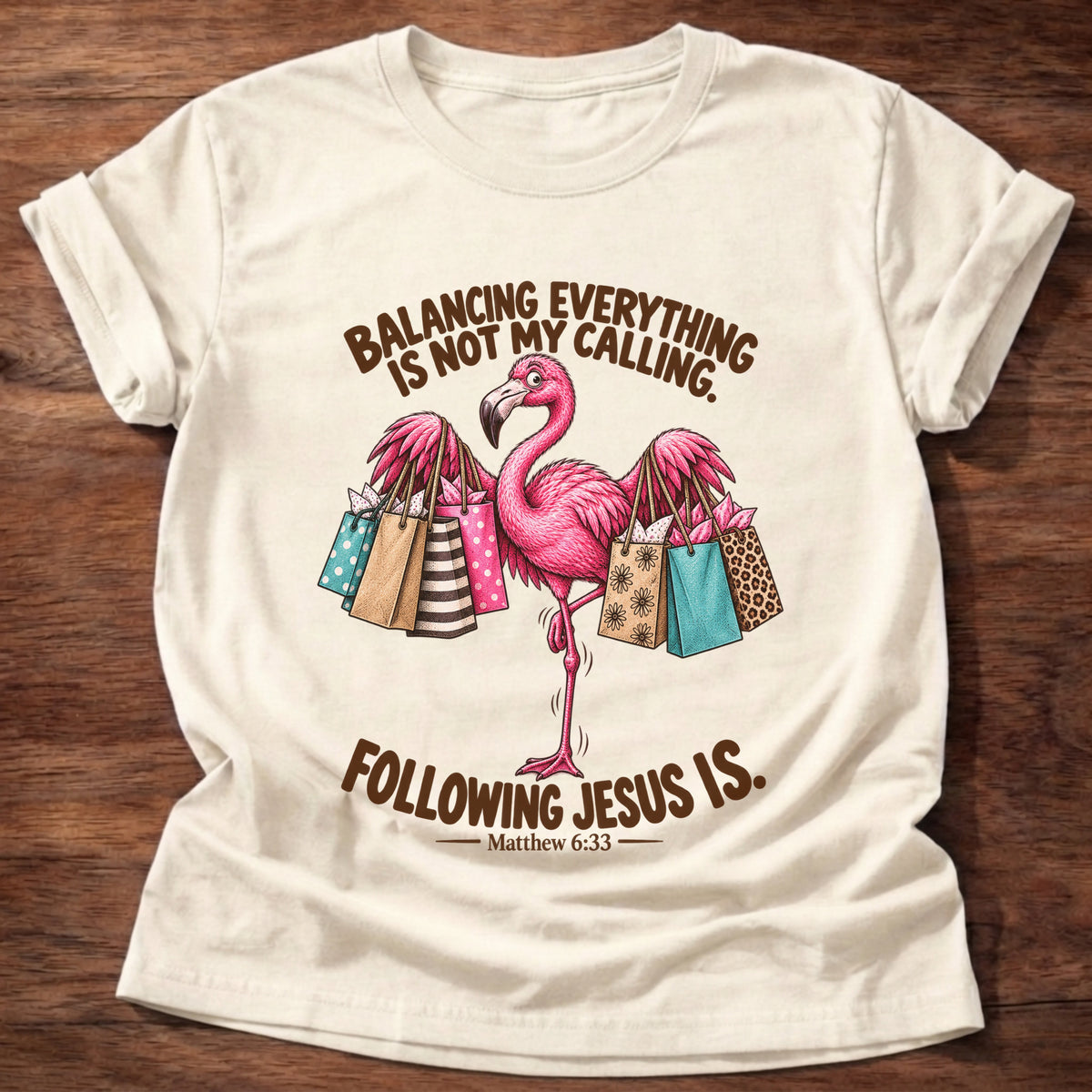 Natural t-shirt printed with a pink flamingo balancing on one leg loaded with colorful shopping bags graphic and “Balancing everything is not my calling. Following Jesus is.” Matthew 6:33 faith based gift