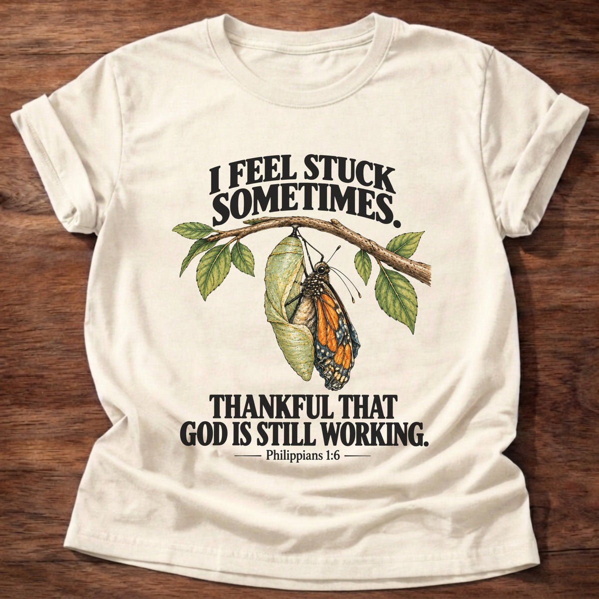 Natural t-shirt printed with a monarch butterfly emerging from its chrysalis on a leafy branch graphic and “I feel stuck sometimes. Thankful that God is still working.” Philippians 1:6 faith based gift