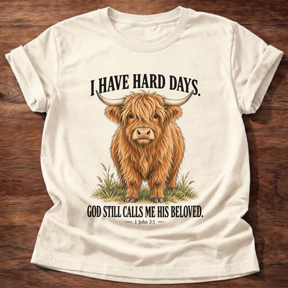 Natural t-shirt printed with a shaggy Highland cow standing in tall grass graphic and “I have hard days. God still calls me His beloved.” 1 John 3:1 faith based gift
