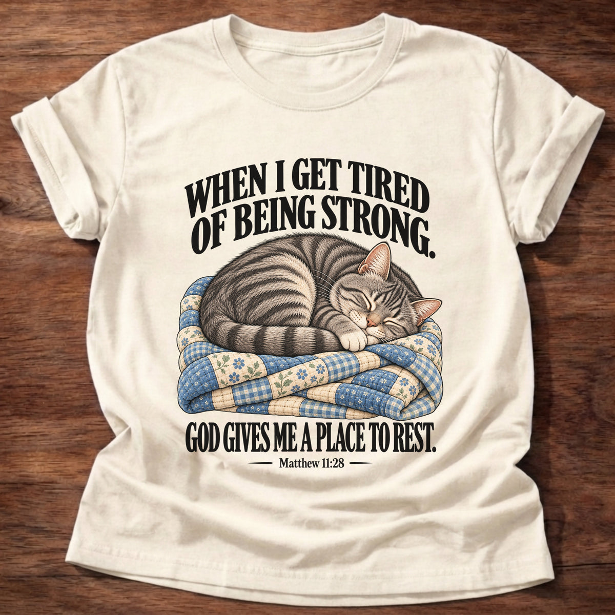 Natural t-shirt printed with a tabby cat curled up asleep on a folded patchwork quilt graphic and “When I get tired of being strong, God gives me a place to rest.” Matthew 11:28 faith based gift