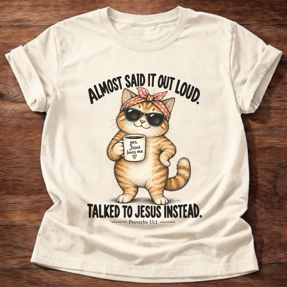 Natural t-shirt printed with an orange tabby cat in sunglasses and a polka-dot bandana holding a 'yes, Jesus loves me' mug graphic and “Almost said it out loud. Talked to Jesus instead.” Proverbs 15:1 faith based gift