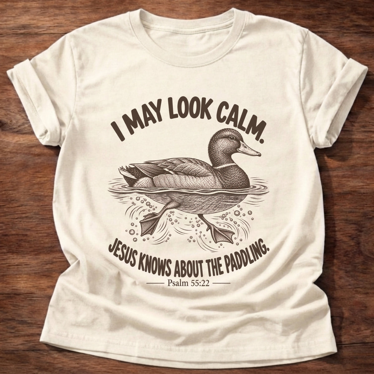 Natural t-shirt printed with a mallard duck gliding on the water with its feet paddling underneath graphic and “I may look calm. Jesus knows about the paddling.” Psalm 55:22 faith based gift