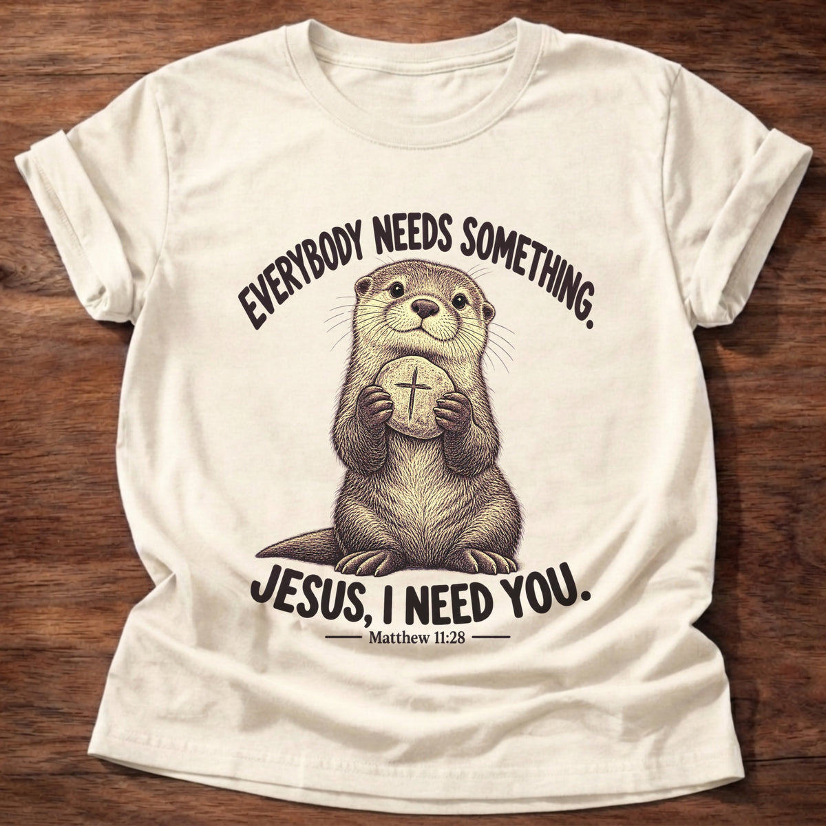 Natural t-shirt printed with an otter hugging a round stone etched with a cross graphic and “Everybody needs something. Jesus, I need you.” Matthew 11:28 faith based gift