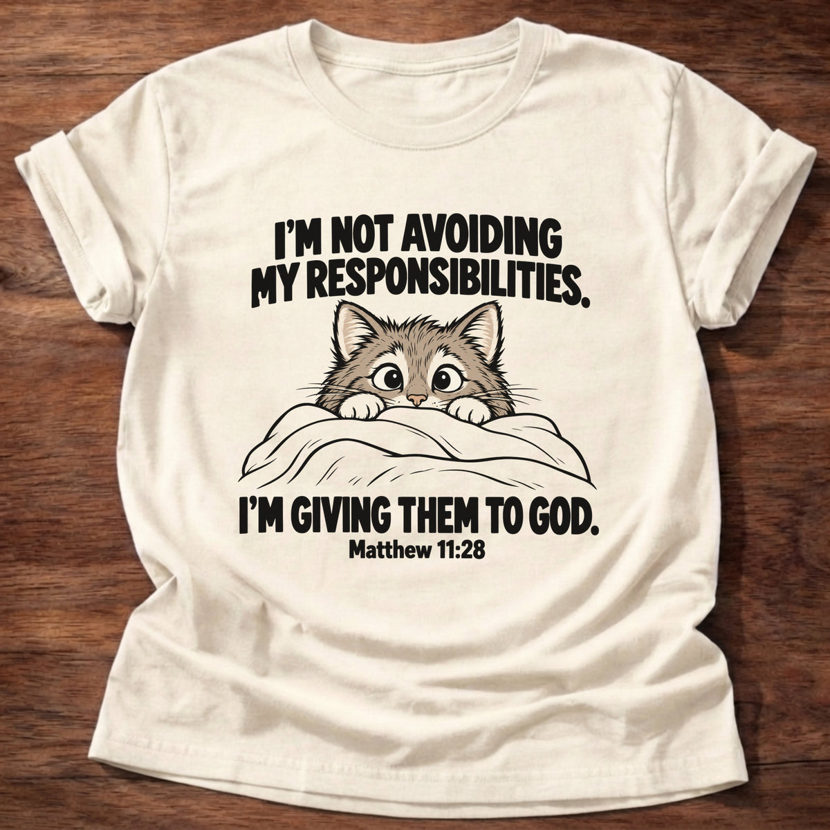 Natural t-shirt printed with a cat peeking over a blanket graphic and 'I'm not avoiding my responsibilities. I'm giving them to God.' Matthew 11:28 faith based gift