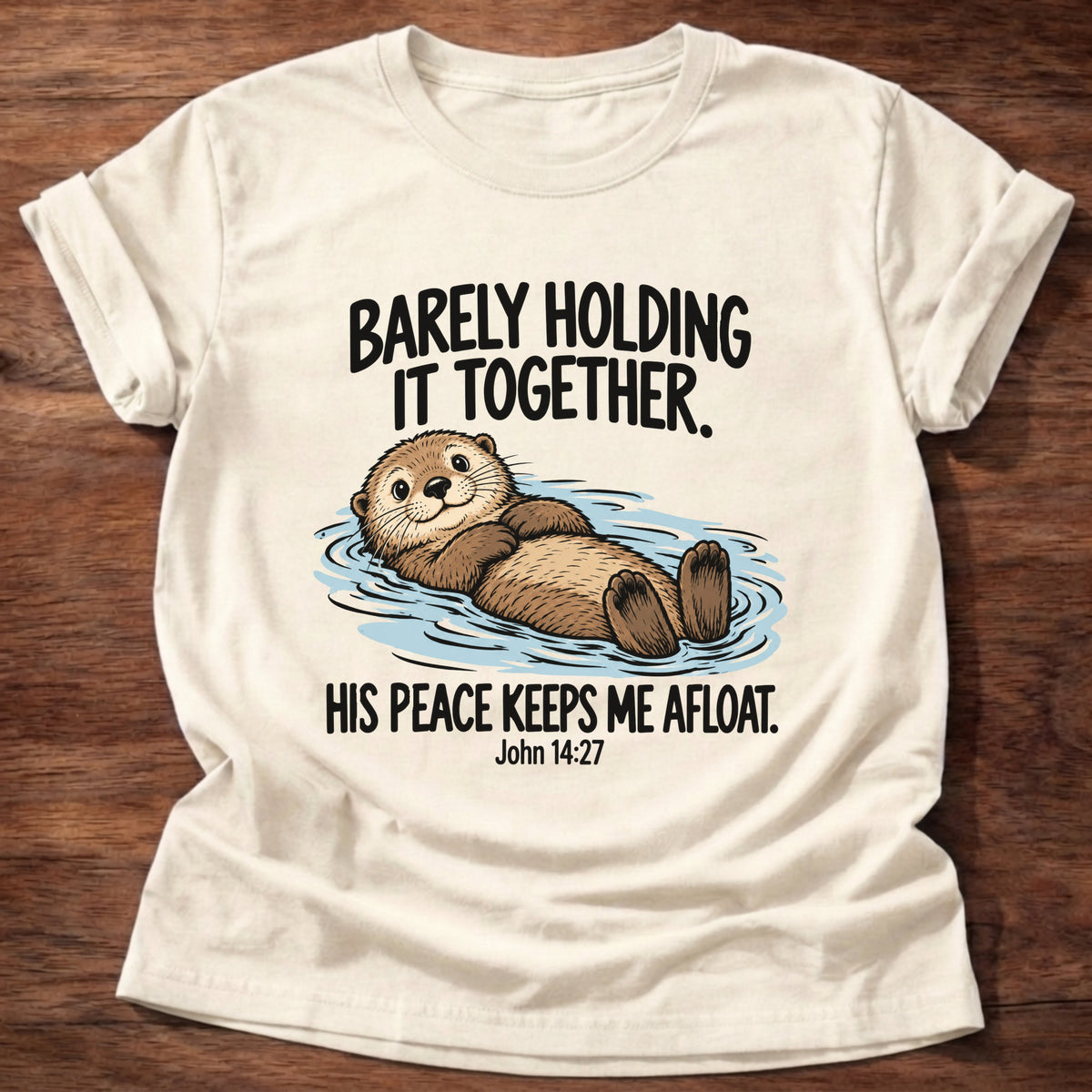 Natural t-shirt printed with an otter floating on its back graphic and 'Barely holding it together. His peace keeps me afloat.' John 14:27 faith based gift
