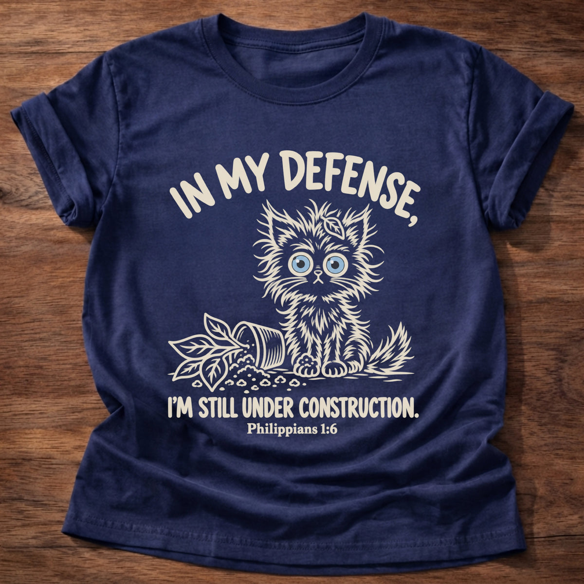 Navy t-shirt printed with a fluffy kitten next to a knocked-over potted plant graphic and “In my defense, I'm still under construction.” Philippians 1:6 Christian shirt