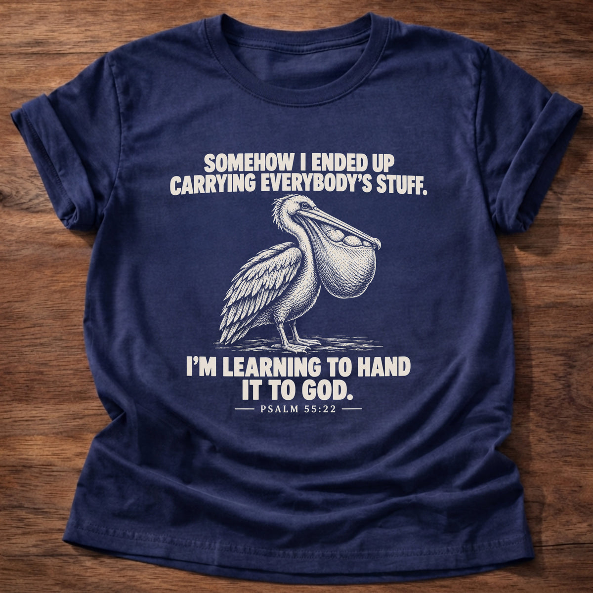 Navy t-shirt printed with a pelican with a pouch full of fish graphic and 'Somehow I ended up carrying everybody's stuff. I'm learning to hand it to God.' Psalm 55:22 faith based gift
