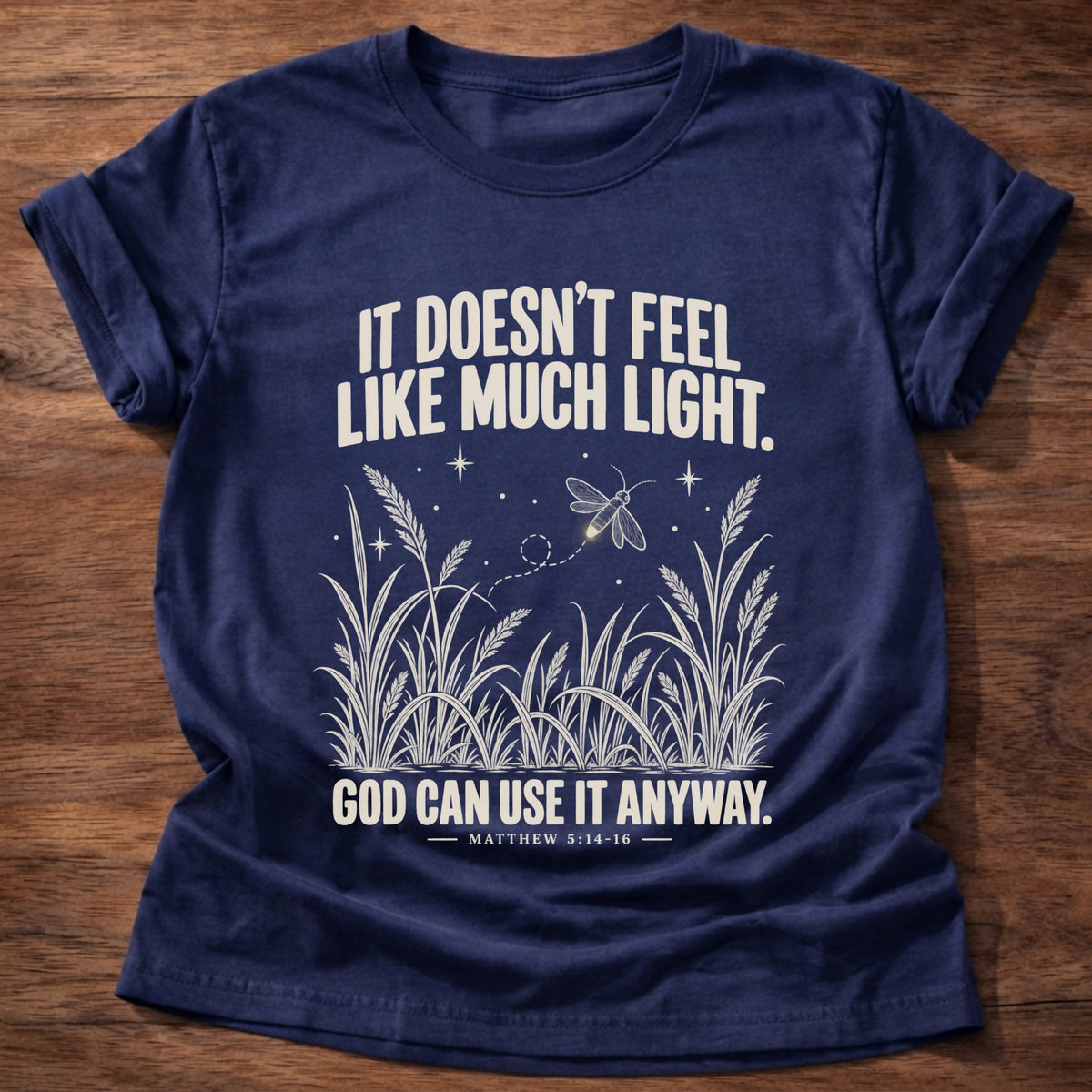 Navy t-shirt printed with a firefly glowing in a dark field of grass graphic and 'It doesn't feel like much light. God can use it anyway.' Matthew 5:14-16 faith based gift