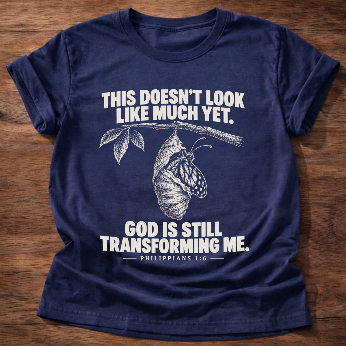 Navy t-shirt printed with a chrysalis hanging from a branch graphic and 'This doesn't look like much yet. God is still transforming me.' Philippians 1:6 faith based gift
