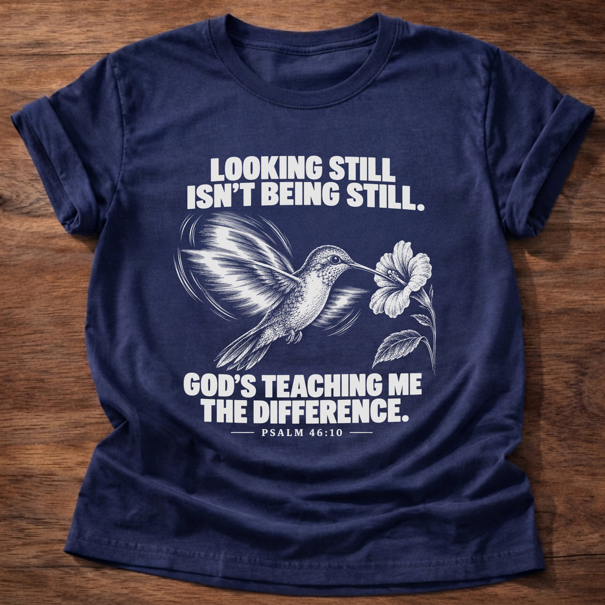 Navy t-shirt printed with a hummingbird hovering beside a flower graphic and 'Looking still isn't being still. God's teaching me the difference.' Psalm 46:10 faith based gift