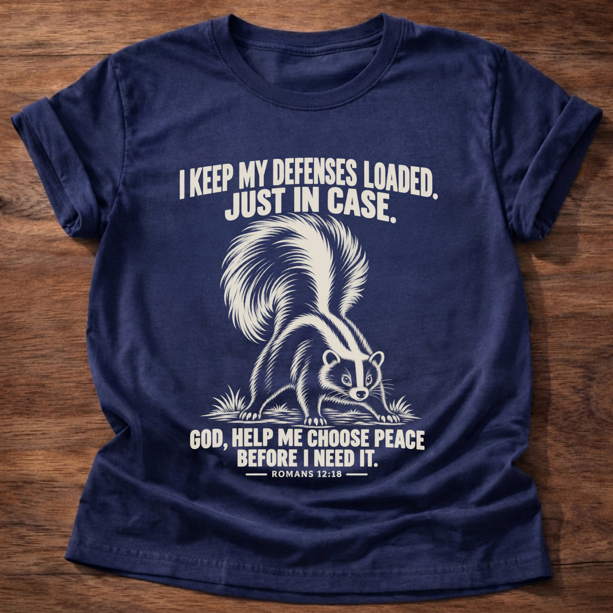 Navy t-shirt printed with a skunk with its tail raised, ready to spray graphic and 'I keep my defenses loaded. Just in case. God, help me choose peace before I need it.' Romans 12:18 faith based gift