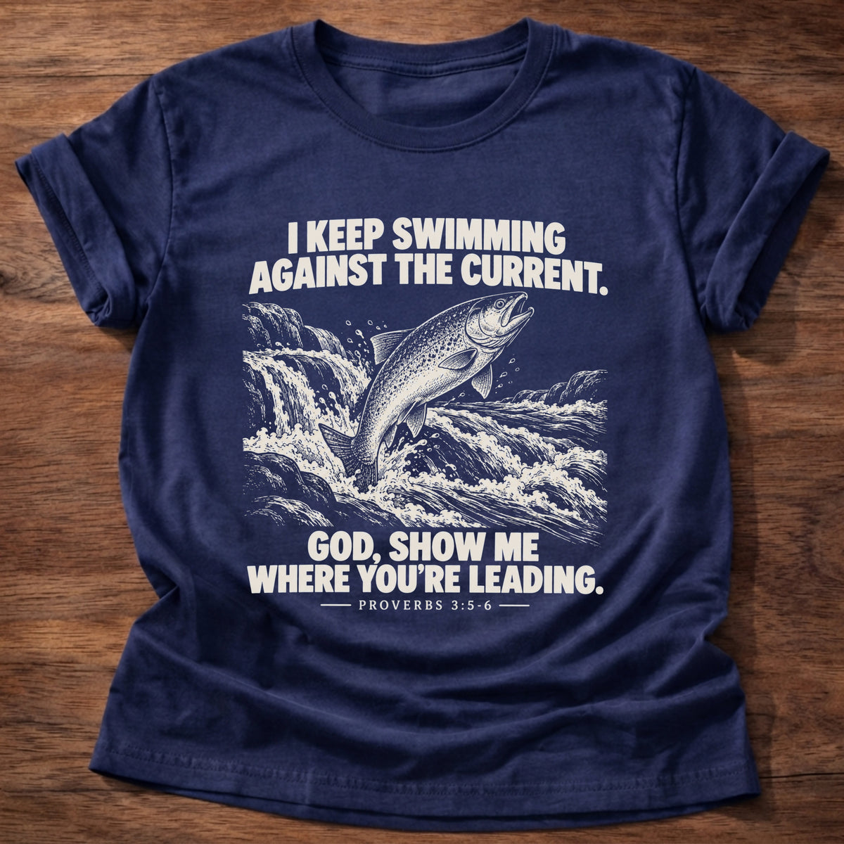 Navy t-shirt printed with a fish leaping against a rushing current graphic and 'I keep swimming against the current. God, show me where you're leading.' Proverbs 3:5-6 faith based gift