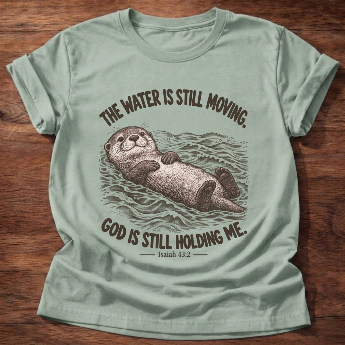 Sage t-shirt printed with an otter floating on its back in moving water graphic and “The water is still moving. God is still holding me.” Isaiah 43:2 faith based gift