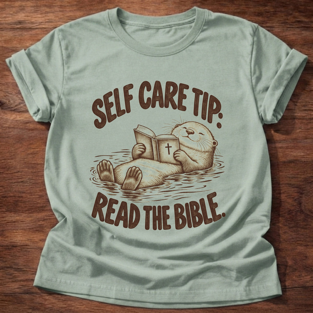 Sage t-shirt printed with a sea otter floating on its back reading a Bible graphic and “Self care tip: Read the Bible.” faith based gift