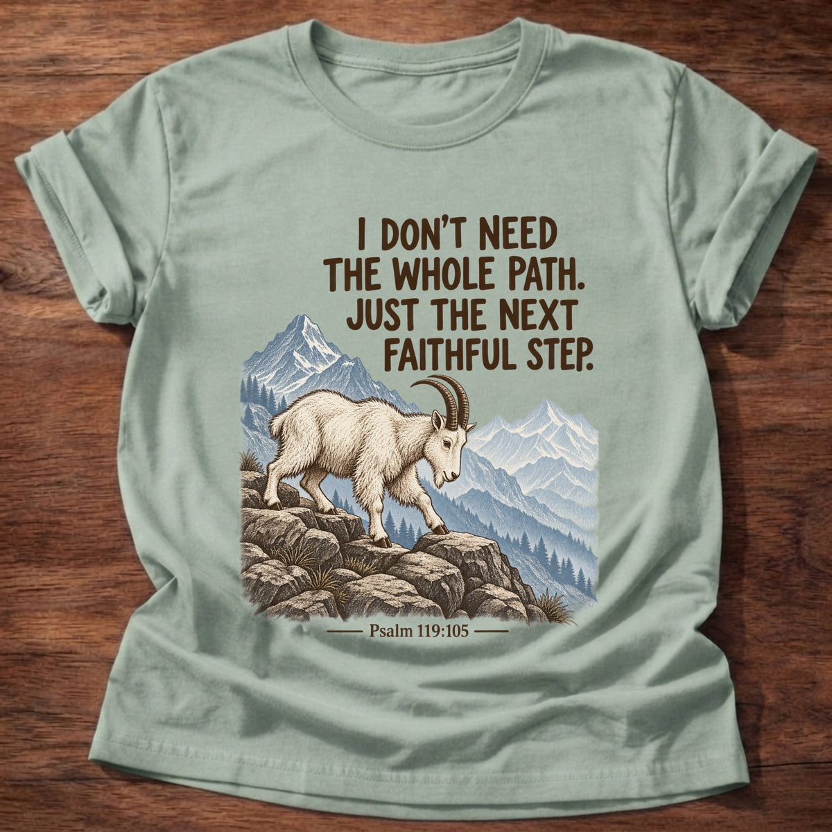 Sage t-shirt printed with a mountain goat stepping across rocky ledges with mountain peaks behind graphic and “I don't need the whole path. Just the next faithful step.” Psalm 119:105 faith based gift