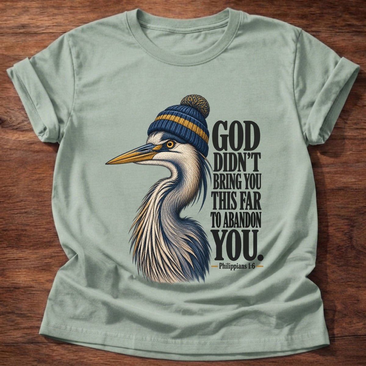 Sage t-shirt printed with a great blue heron wearing a knit beanie graphic and “God didn't bring you this far to abandon you.” Philippians 1:6 faith based gift