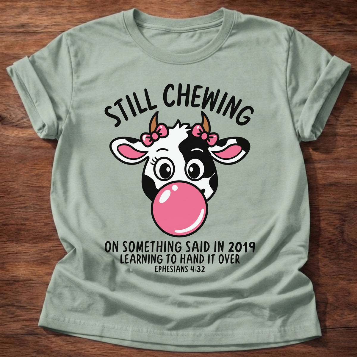 Sage t-shirt printed with a cow blowing a bubblegum bubble and “Still chewing on something said in 2019. Learning to hand it over.” Ephesians 4:32 Christian shirt