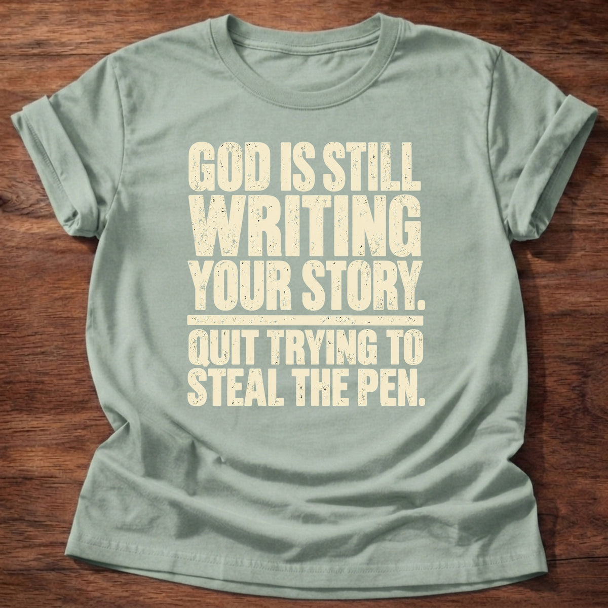 Sage t-shirt printed with bold stacked typography graphic and “God is still writing your story. Quit trying to steal the pen.” Philippians 1:6 Christian shirt