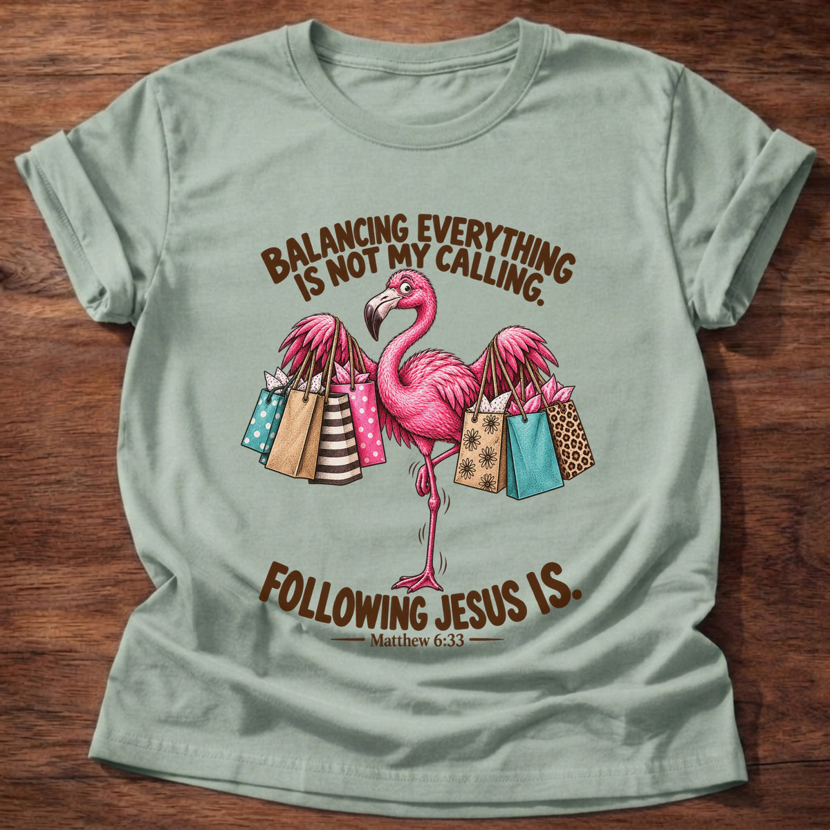 Sage t-shirt printed with a pink flamingo balancing on one leg loaded with colorful shopping bags graphic and “Balancing everything is not my calling. Following Jesus is.” Matthew 6:33 faith based gift