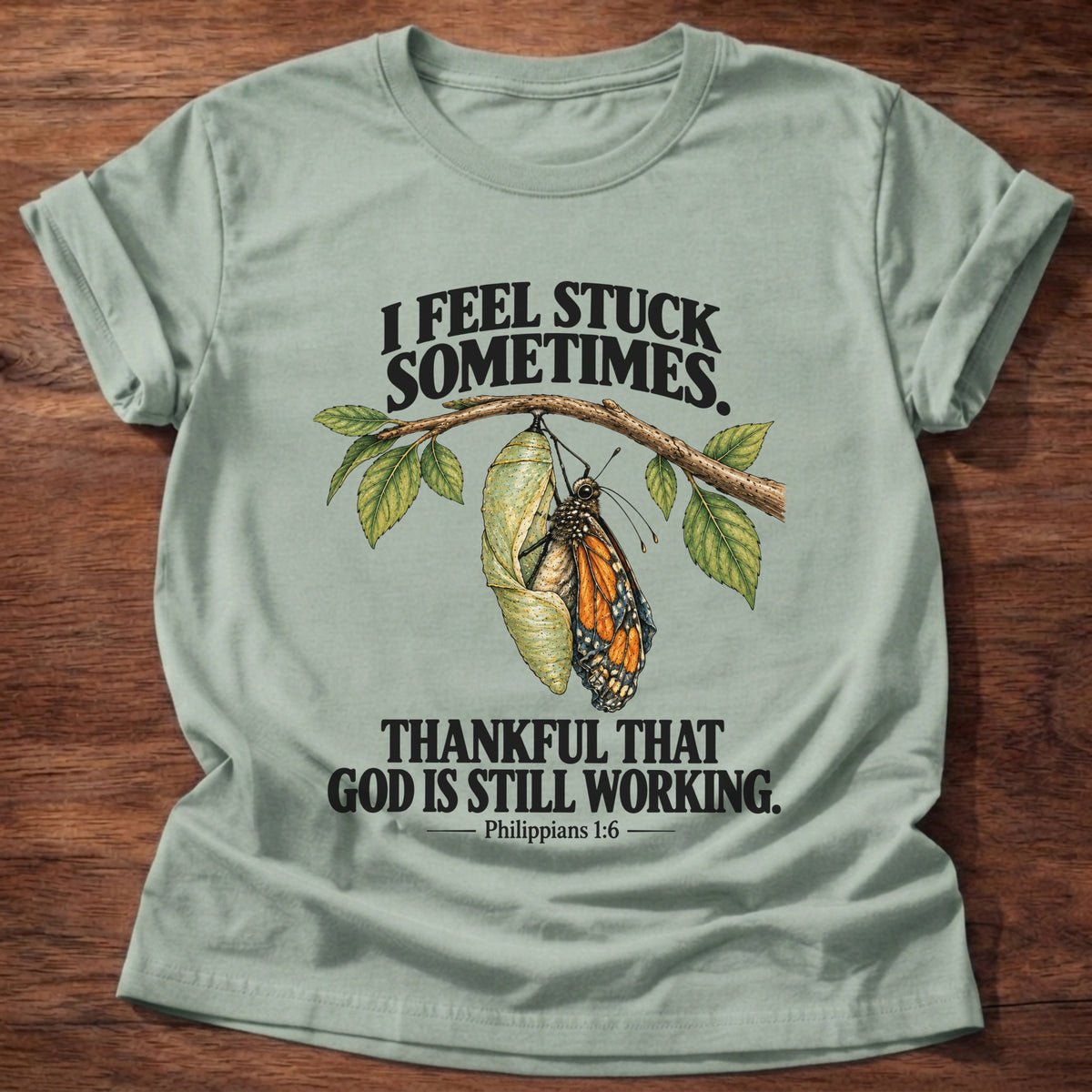 Sage t-shirt printed with a monarch butterfly emerging from its chrysalis on a leafy branch graphic and “I feel stuck sometimes. Thankful that God is still working.” Philippians 1:6 faith based gift