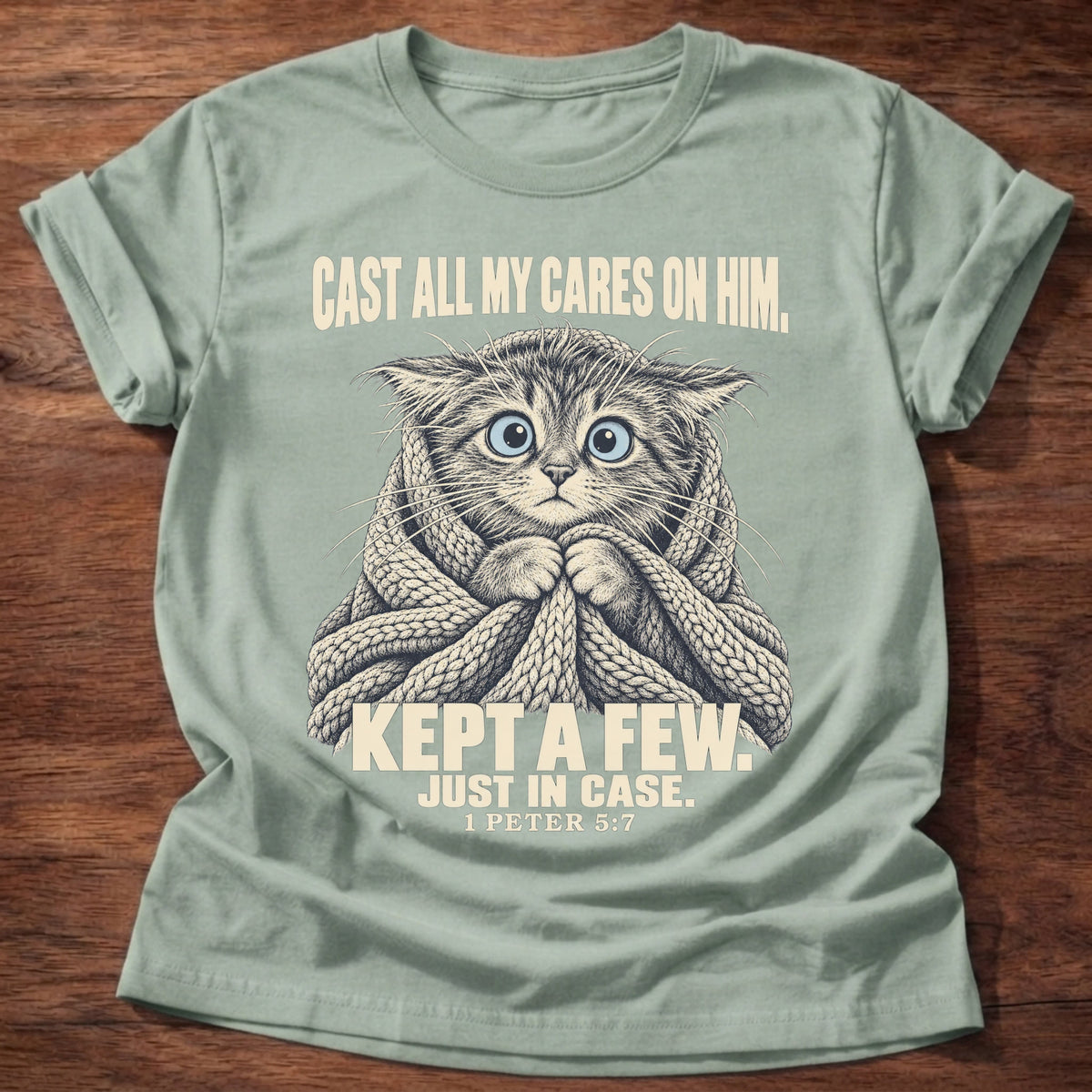 Kept A Few T-Shirt in Sage featuring a wide-eyed cat wrapped in a knit blanket and 1 Peter 5:7 anxiety humor design