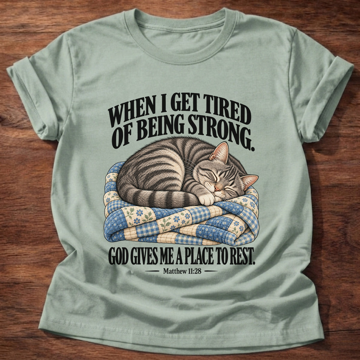 Sage t-shirt printed with a tabby cat curled up asleep on a folded patchwork quilt graphic and “When I get tired of being strong, God gives me a place to rest.” Matthew 11:28 faith based gift