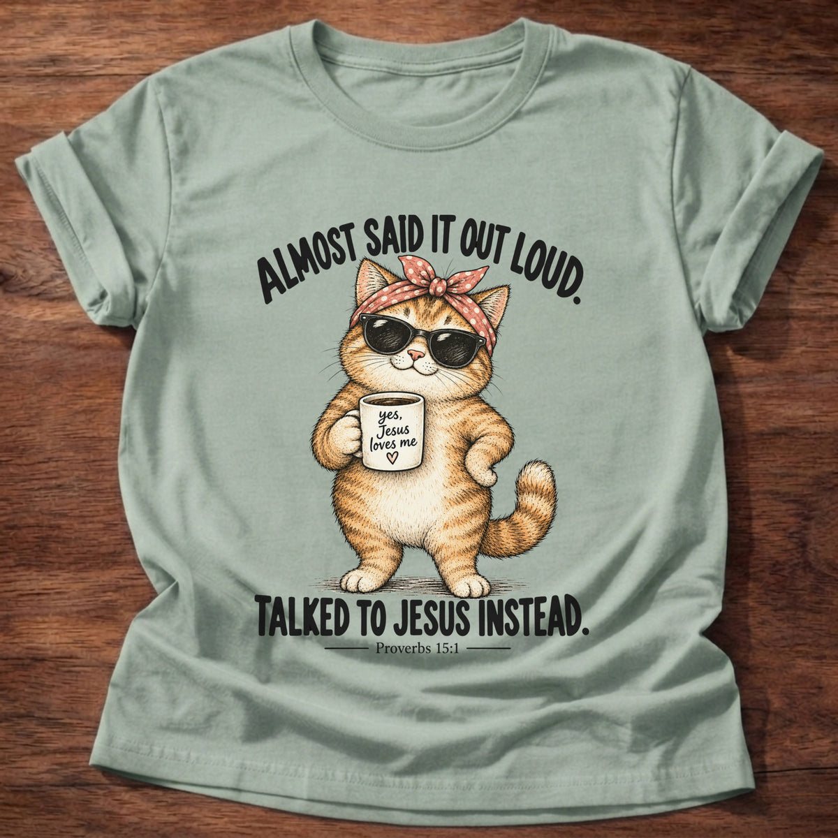 Sage t-shirt printed with an orange tabby cat in sunglasses and a polka-dot bandana holding a 'yes, Jesus loves me' mug graphic and “Almost said it out loud. Talked to Jesus instead.” Proverbs 15:1 faith based gift