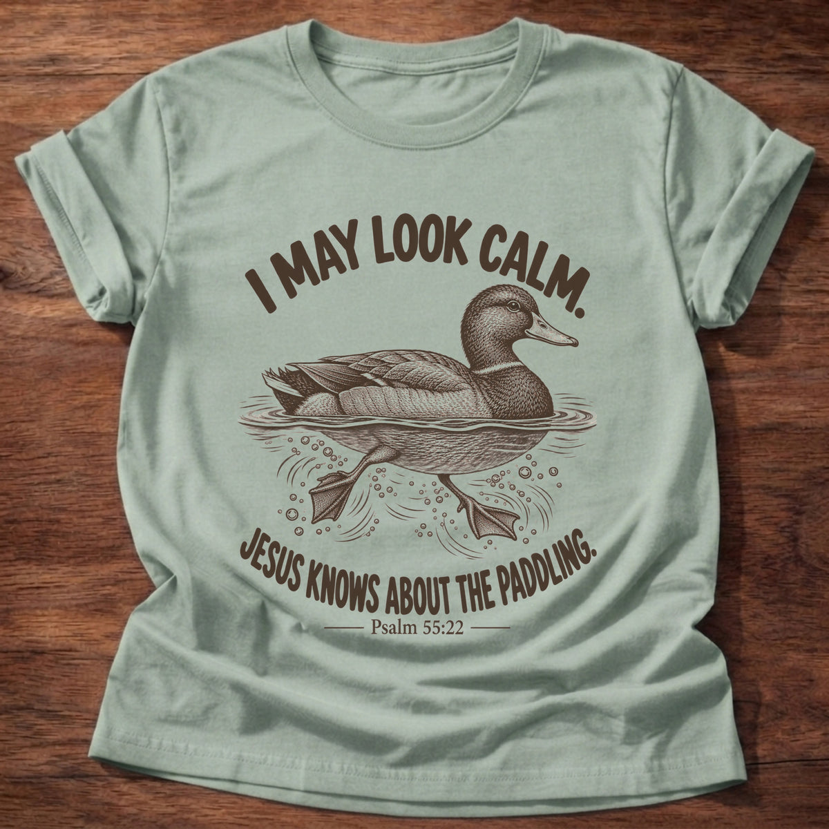 Sage t-shirt printed with a mallard duck gliding on the water with its feet paddling underneath graphic and “I may look calm. Jesus knows about the paddling.” Psalm 55:22 faith based gift