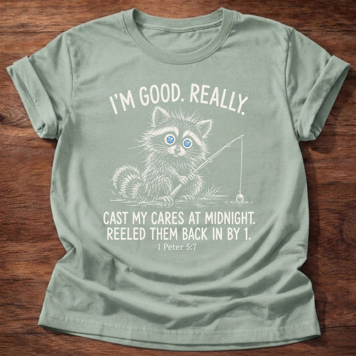 Sage t-shirt printed with a raccoon holding a fishing rod graphic and “I'm good. Really. Cast my cares at midnight. Reeled them back in by 1.” 1 Peter 5:7 Christian shirt