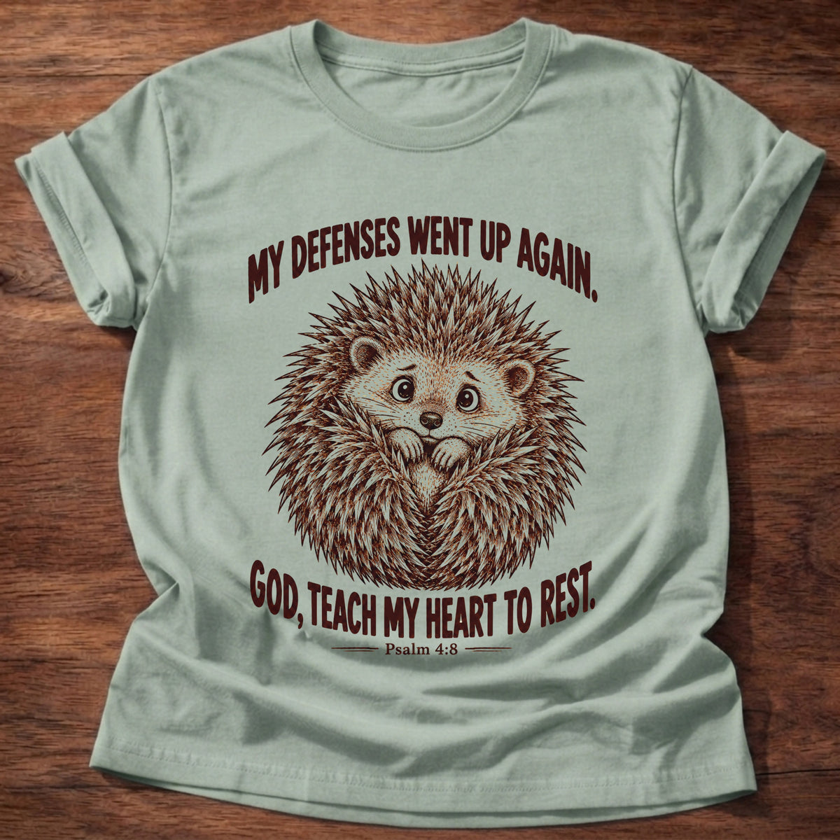 Sage t-shirt printed with a worried hedgehog curled up with its quills raised graphic and “My defenses went up again. God, teach my heart to rest.” Psalm 4:8 faith based gift