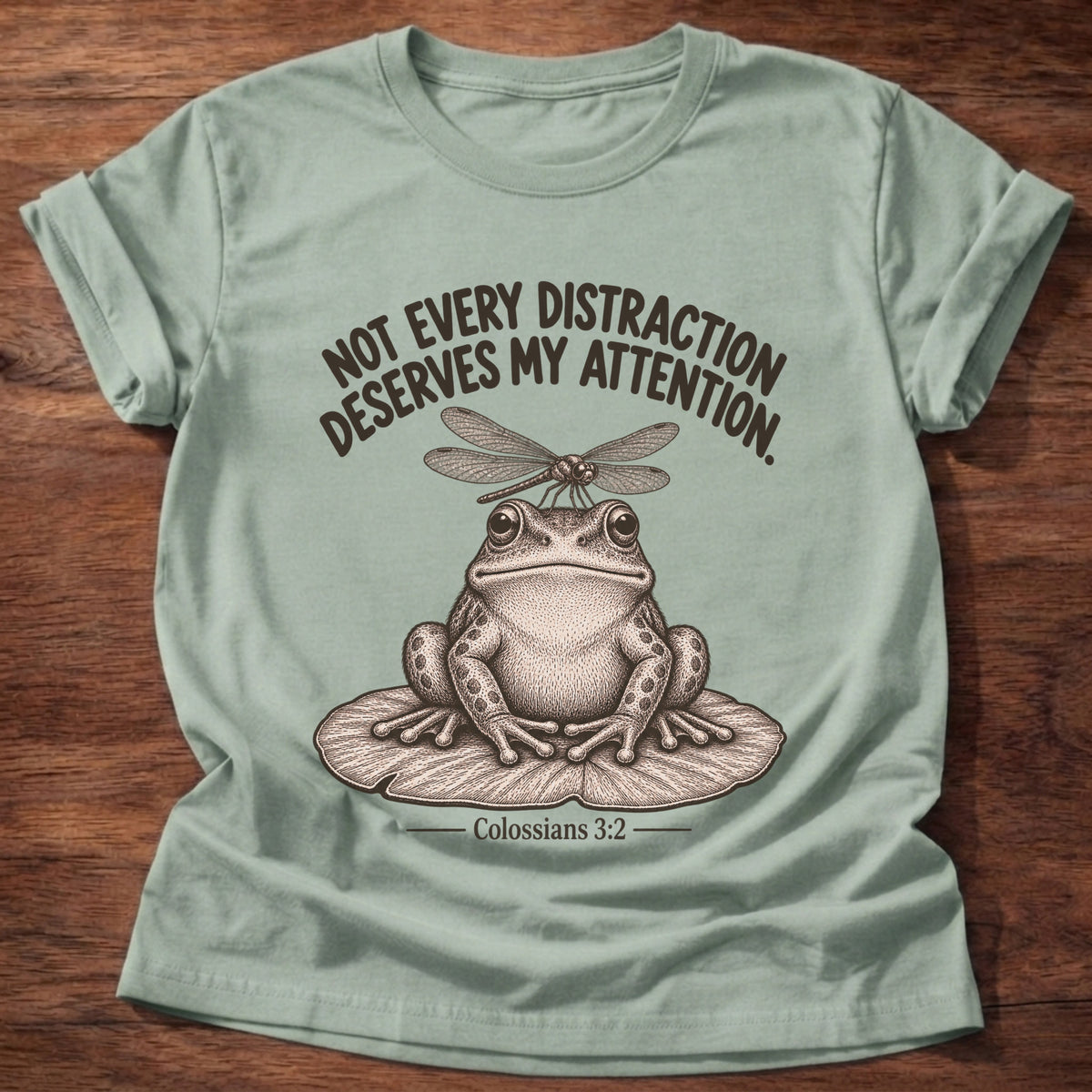 Sage t-shirt printed with a frog sitting on a lily pad with a dragonfly perched on its head graphic and “Not every distraction deserves my attention.” Colossians 3:2 faith based gift