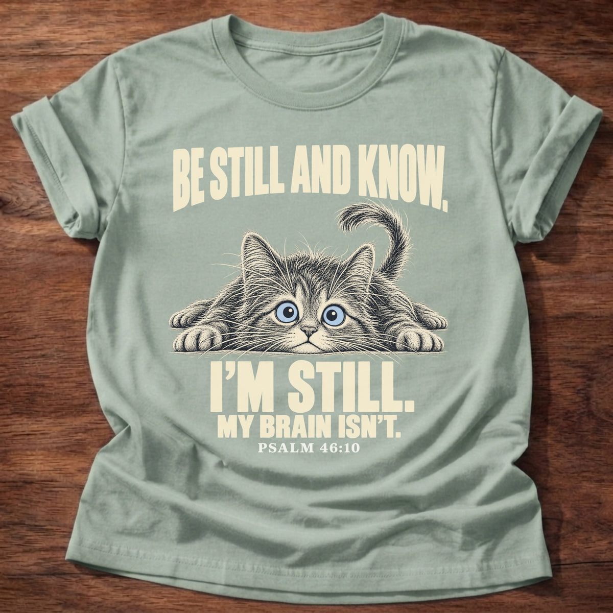 Brain Not Still T-Shirt in Sage featuring a wide-eyed cat lying flat with alert eyes and Psalm 46:10 overthinking humor design