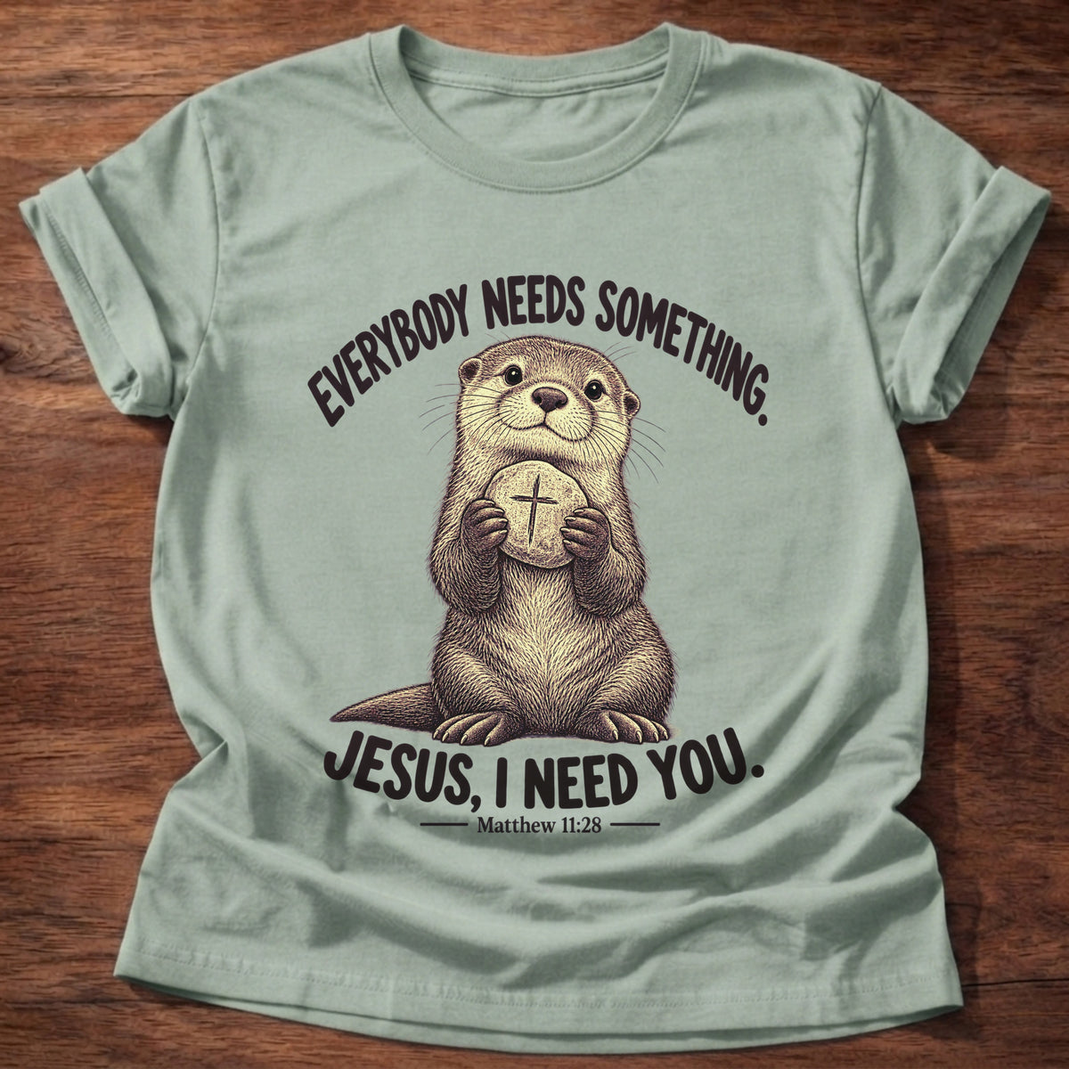 Sage t-shirt printed with an otter hugging a round stone etched with a cross graphic and “Everybody needs something. Jesus, I need you.” Matthew 11:28 faith based gift