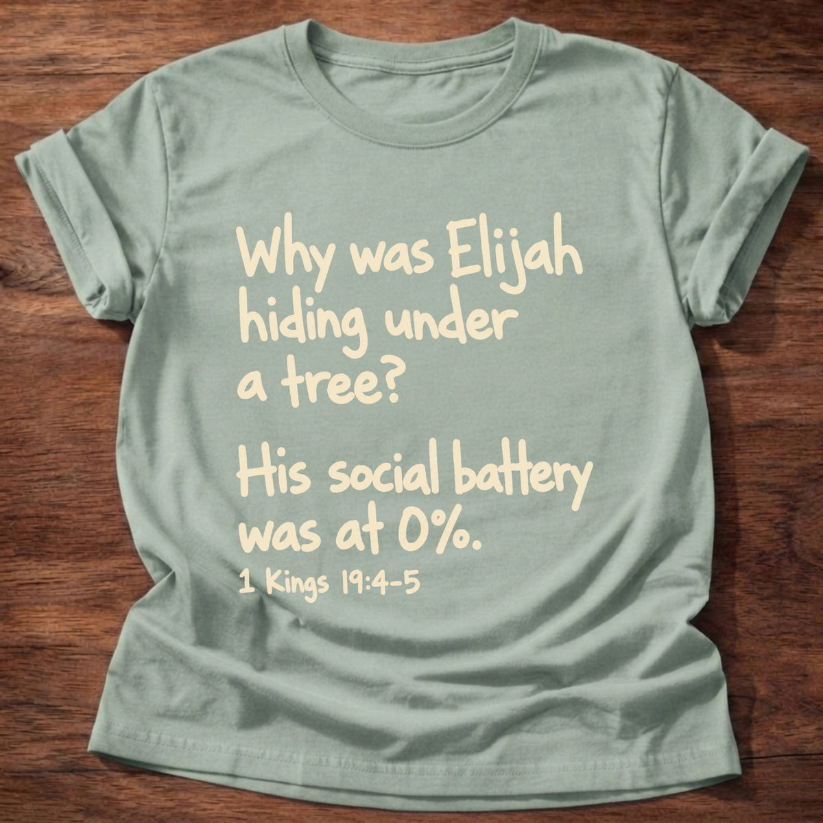 Sage t-shirt printed with a simple hand-lettered Bible joke graphic and “Why was Elijah hiding under a tree? His social battery was at 0%.” 1 Kings 19:4-5 Christian shirt