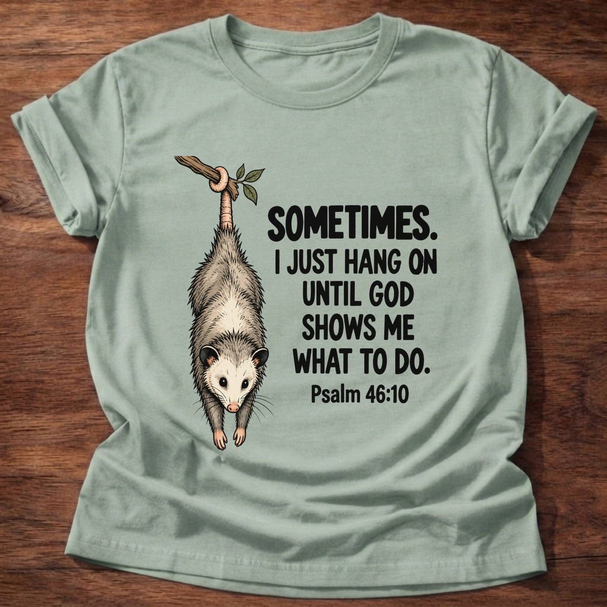 Sage t-shirt printed with a possum hanging by its tail graphic and 'Sometimes. I just hang on until God shows me what to do.' Psalm 46:10 faith based gift
