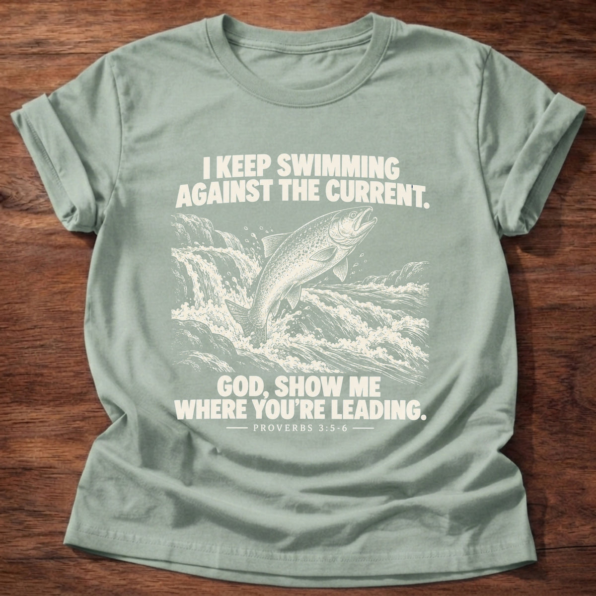 Sage t-shirt printed with a fish leaping against a rushing current graphic and 'I keep swimming against the current. God, show me where you're leading.' Proverbs 3:5-6 faith based gift