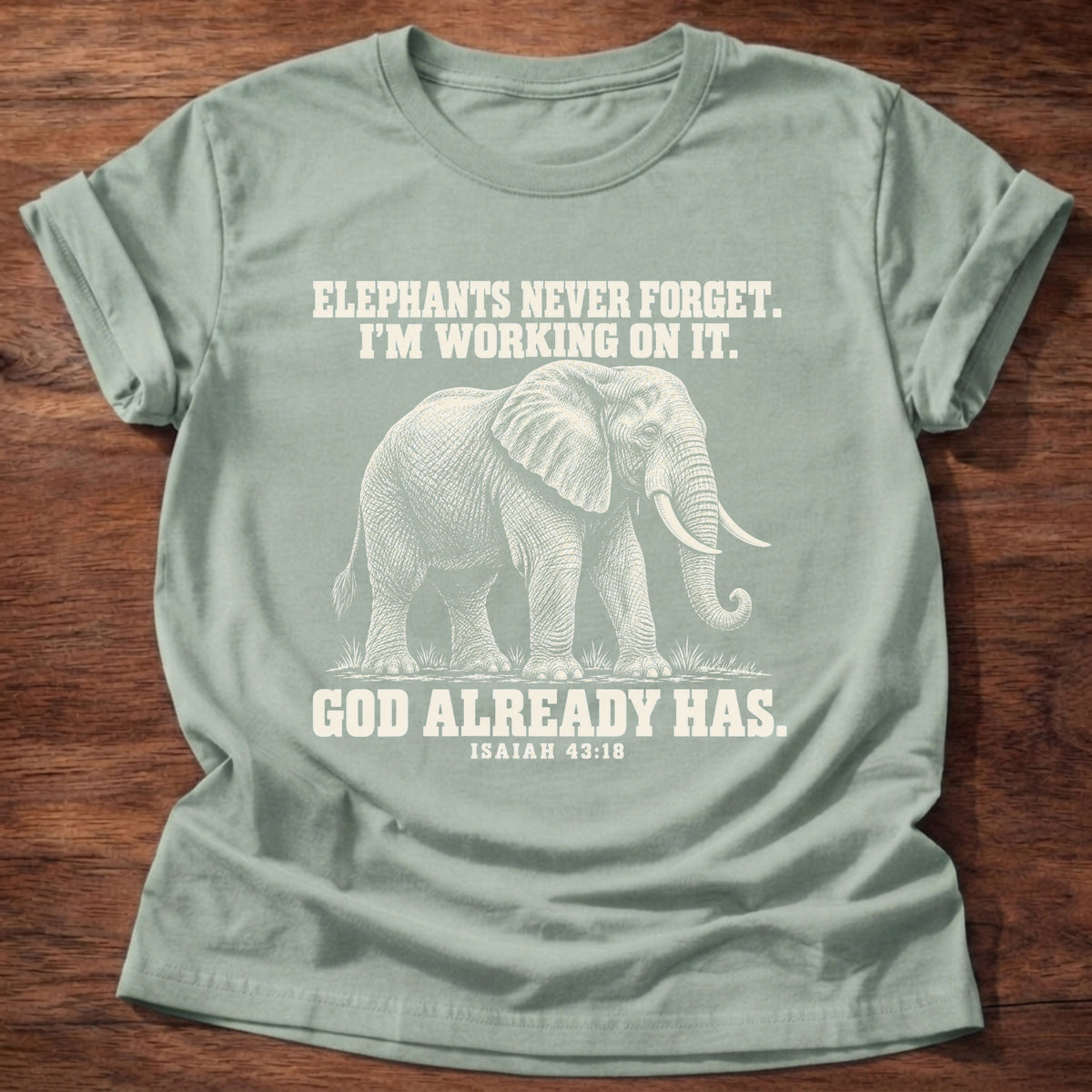 Sage t-shirt printed with an elephant walking through tall grass graphic and 'Elephants never forget. I'm working on it. God already has.' Isaiah 43:18 faith based gift