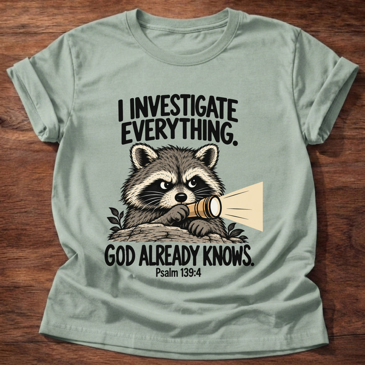 Sage t-shirt printed with a raccoon holding a magnifying glass graphic and 'I investigate everything. God already knows.' Psalm 139:4 faith based gift