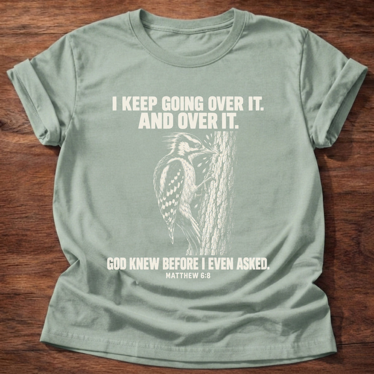 Sage t-shirt printed with a woodpecker drumming the same spot on a tree graphic and 'I keep going over it. And over it. God knew before I even asked.' Matthew 6:8 faith based gift