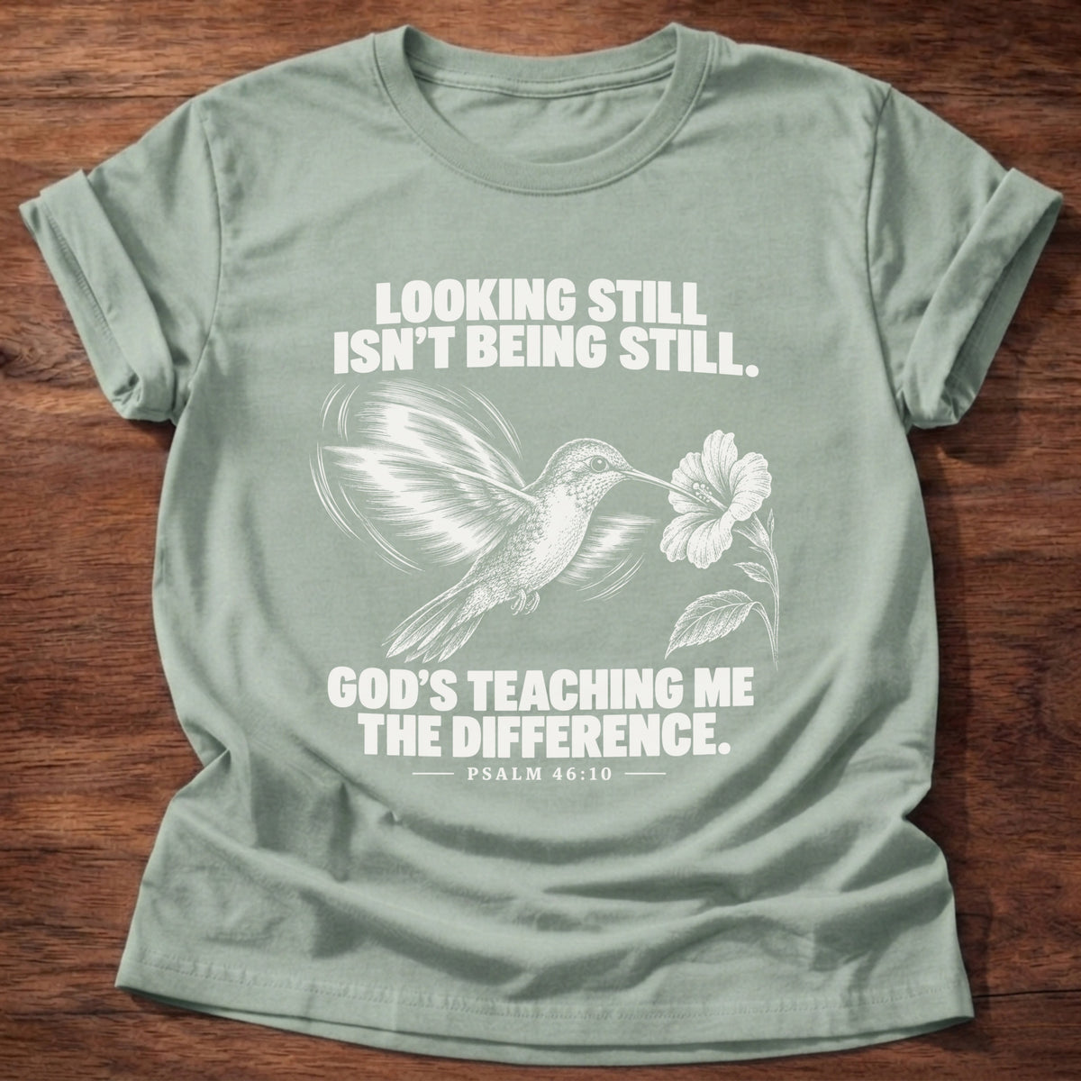 Sage t-shirt printed with a hummingbird hovering beside a flower graphic and 'Looking still isn't being still. God's teaching me the difference.' Psalm 46:10 faith based gift