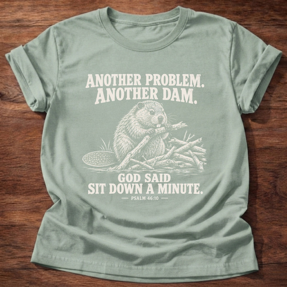 Sage t-shirt printed with a beaver mid-build with another branch graphic and 'Another problem. Another dam. God said sit down a minute.' Psalm 46:10 faith based gift
