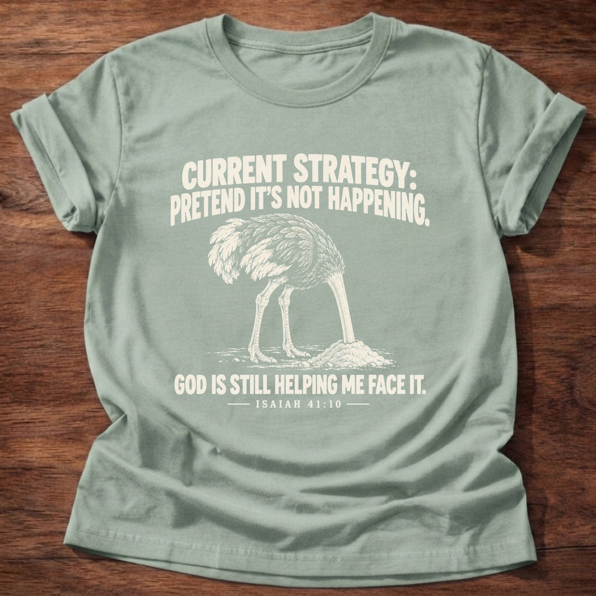 Sage t-shirt printed with an ostrich with its head buried in the sand graphic and 'Current strategy: pretend it's not happening. God is still helping me face it.' Isaiah 41:10 faith based gift