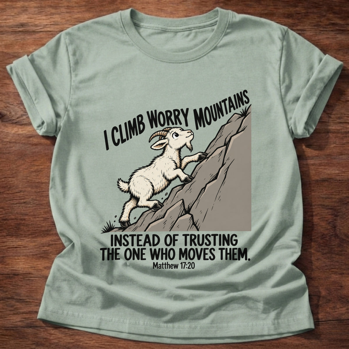 Sage t-shirt printed with a goat climbing a mountain graphic and 'I climb worry mountains instead of trusting the One who moves them.' Matthew 17:20 faith based gift