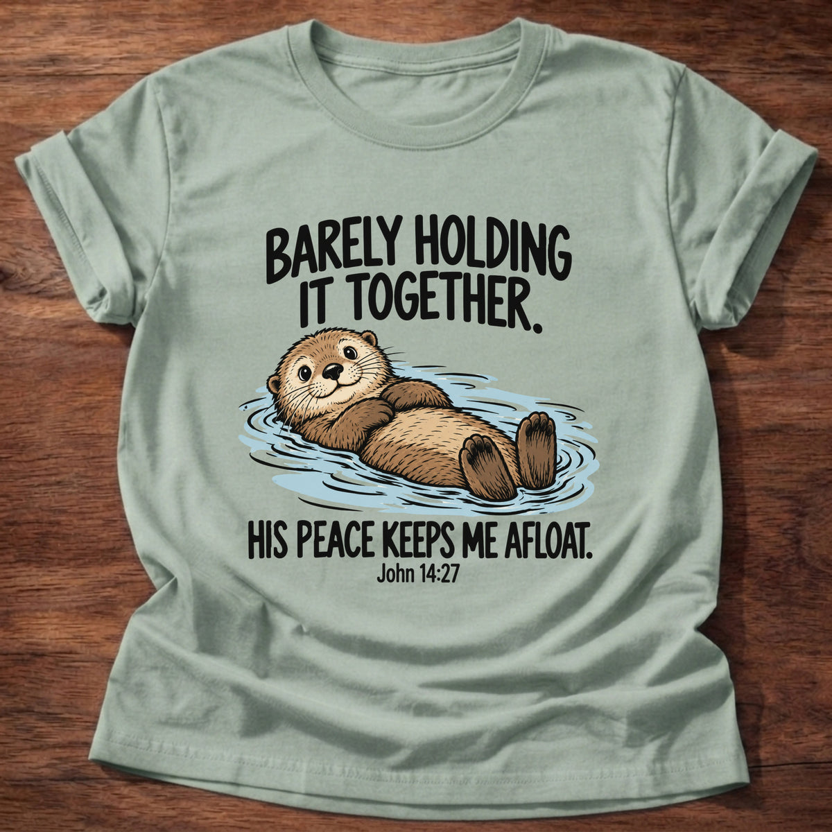Sage t-shirt printed with an otter floating on its back graphic and 'Barely holding it together. His peace keeps me afloat.' John 14:27 faith based gift